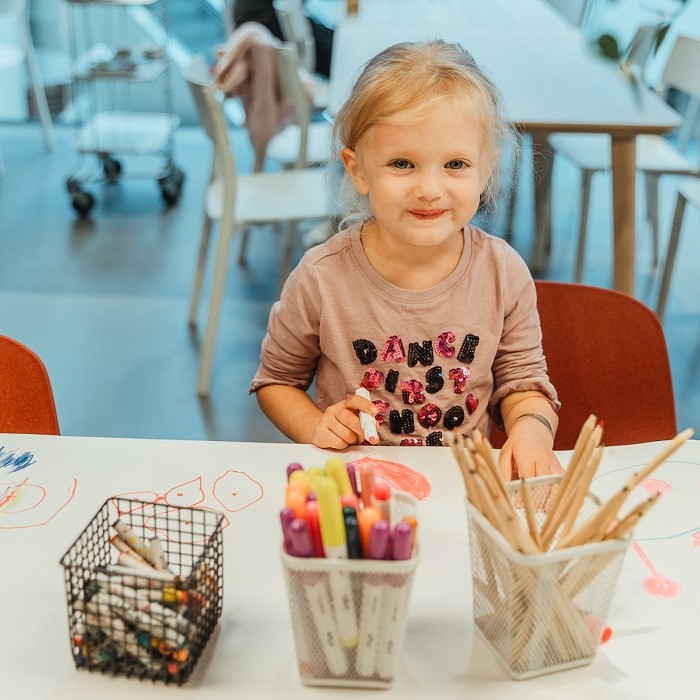 Maza meitene sēž pie galda IKEA veikalā un zīmē ar krāsainiem zīmuļiem. Viņas priekšā ir zīmuļu trauks un rozā papīra sirsniņas.