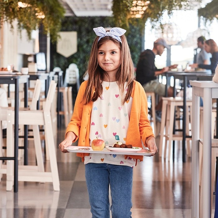 A small girl wearing an orange cardigan and a bow walks through the Swedish restaurant carrying a dish tray.				