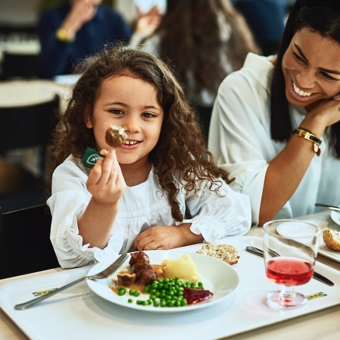 A little girl enjoys a serving of meatballs at a Swedish restaurant. Her smiling mother sits beside her.				