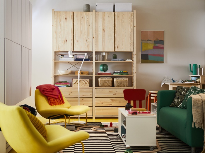 Svetainėje stovi natūralaus medžio daiktų laikymo sistema, žalia sofa, geltonas fotelis su raudonu užtiesalu, baltas kavos staliukas, patiestas dryžuotas kilimas, o ant sienos kabo spalvingas paveikslas. Baldai ir detalės kuria šiltą, jaukią atmosferą.