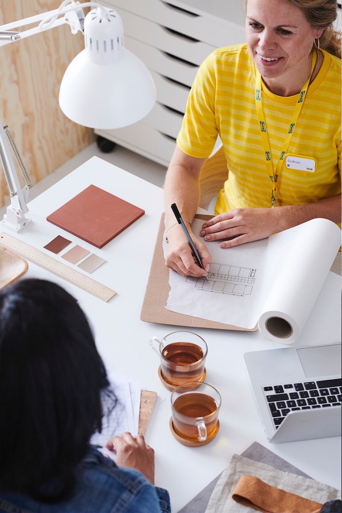 IKEA interjero dizainerė su geltona uniforma konsultacijos metu piešia eskizus ant didelio popieriaus ritinio.