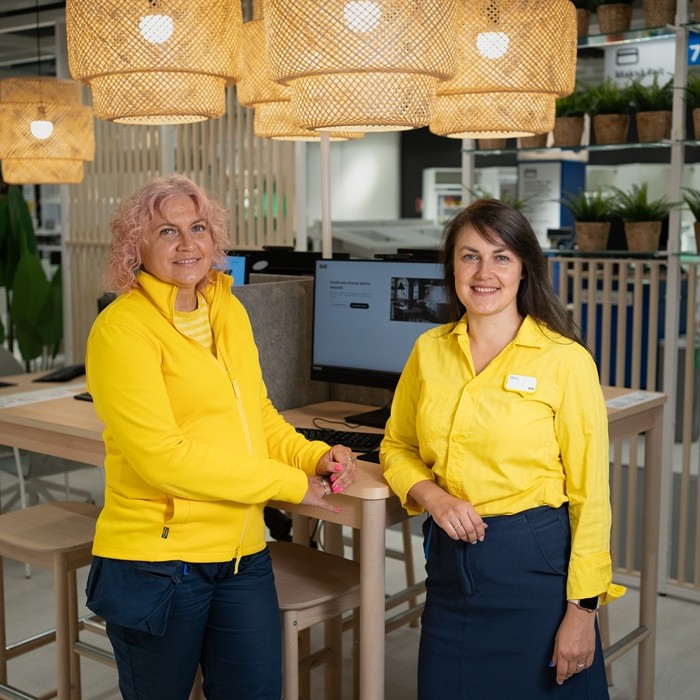 Divas IKEA ekspertes pie galda veikalā ar tumši pelēkiem GLADELIG traukiem uz pīta paliktņa.