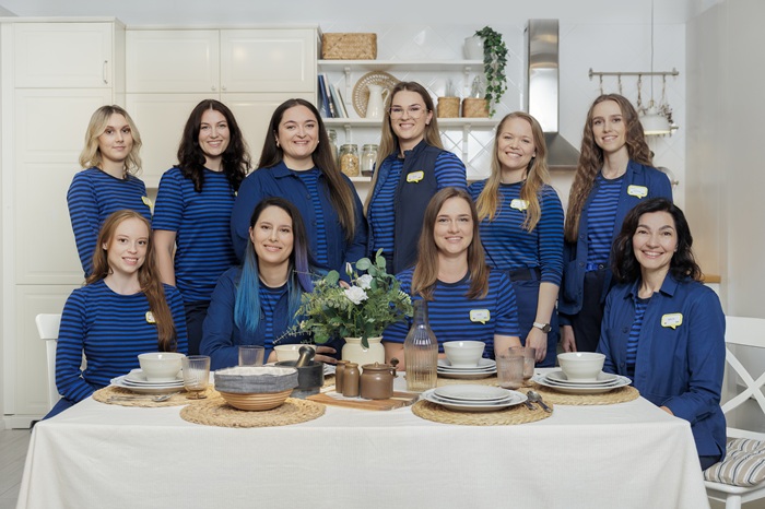 Group of ten interior designers in blue striped uniforms, posing in a modern kitchen setting, around a dining table set with tableware and decor.