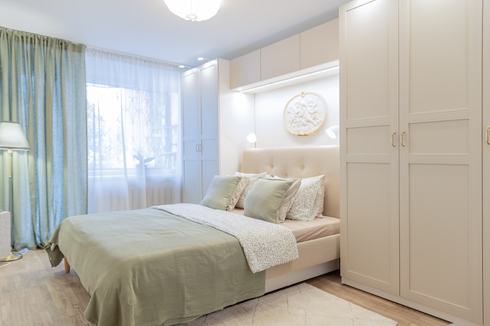 Bright bedroom with a beige upholstered bed, green bedding, white wardrobe and sheer green curtains.
