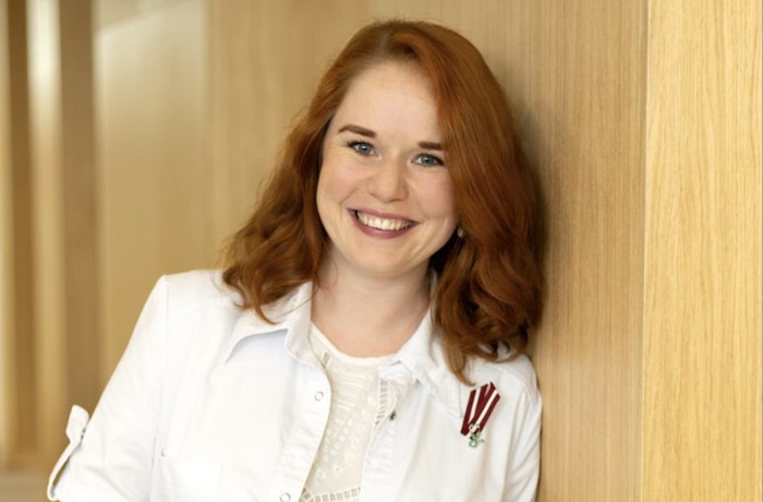 A smiling woman with red hair, dressed in white and wearing a Latvian flag pin, leans against a wooden door.