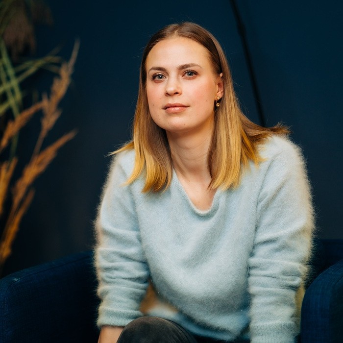 A woman in a light blue sweater sits on a dark blue chair, with dark grasses visible in a flower pot against a dark background.