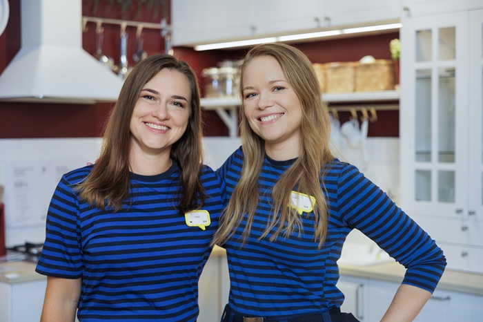 Dvi IKEA interjero dizainerės su mėlynos spalvos uniformomis stovi virtuvėje ir šypsosi.