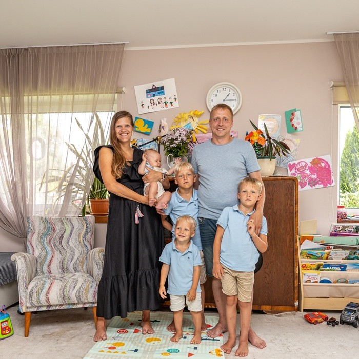 A happy family – 2 parents and 4 kids – stand in a bright living room full of toys and colorful drawings.