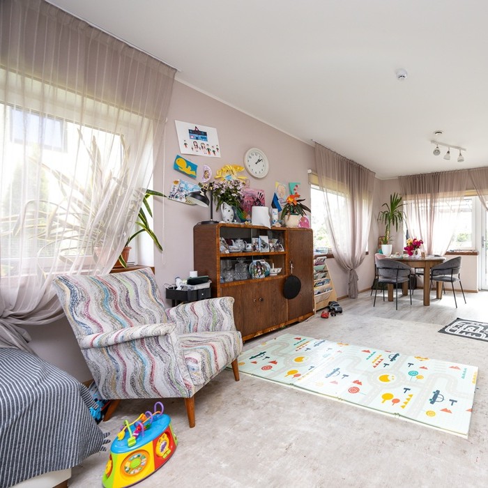 Big and airy living room full of mismatched items of furniture, toys and children´s drawings. There are armchairs on the foreground and a dining area on the background of the picture. 
