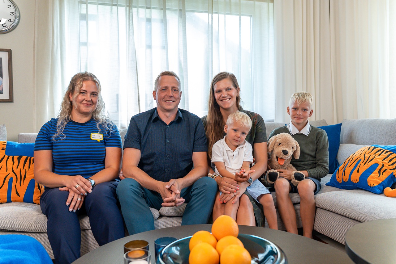 Cheerful IKEA designer sits next to the smiling parents of the big family with 2 of their children on the grey sofa in the newly furnished living room.  