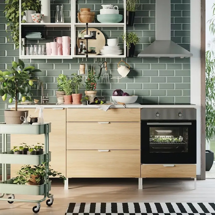 A modern ENHET kitchen with light wood cabinet fronts, a black oven and a green tiled backsplash, tastefully adorned with plants.