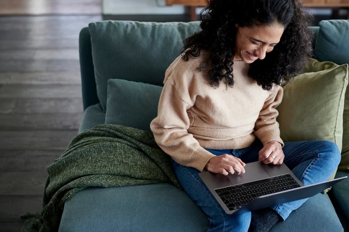 Person with dark curly hair is sitting on a sofa and smiling while using a laptop.