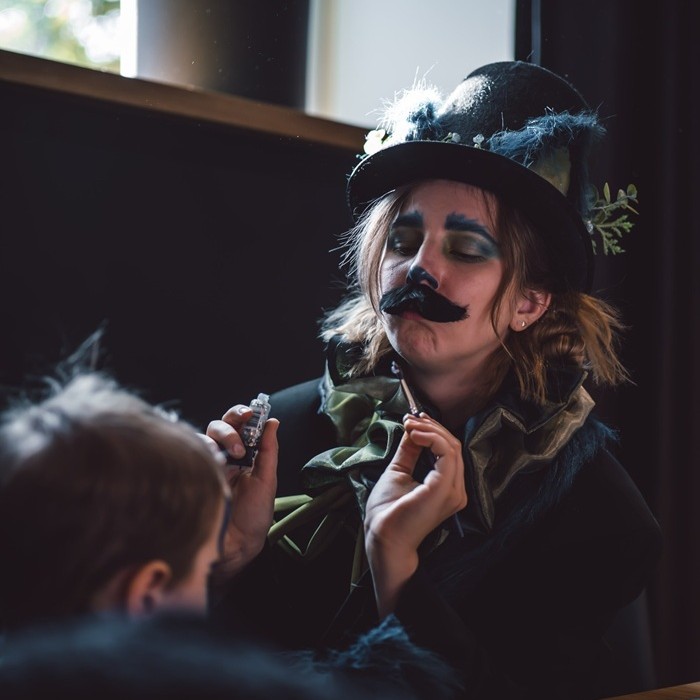 A person dressed in a spooky cat costume with dramatic face paint, including a drawn-on mustache and blue eye shadow, is painting a child’s face.