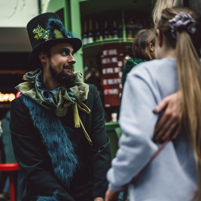 A person in a spooky cat costume with a furry collar, top hat, and face paint smiles while interacting with a child.