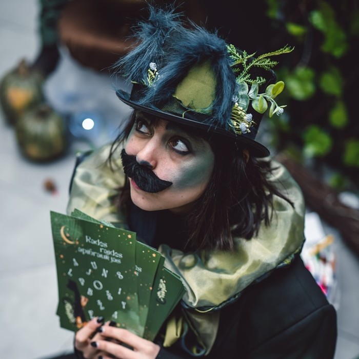 A person in makeup and a spooky cat costume holds green cards with symbols, looking up with a playful expression.