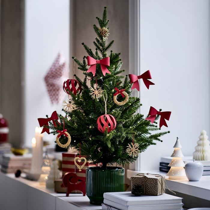 Uz galda stāv Ziemassvētku eglīte podiņā, rotāta ar sarkaniem dažādas formas rotājumiem.