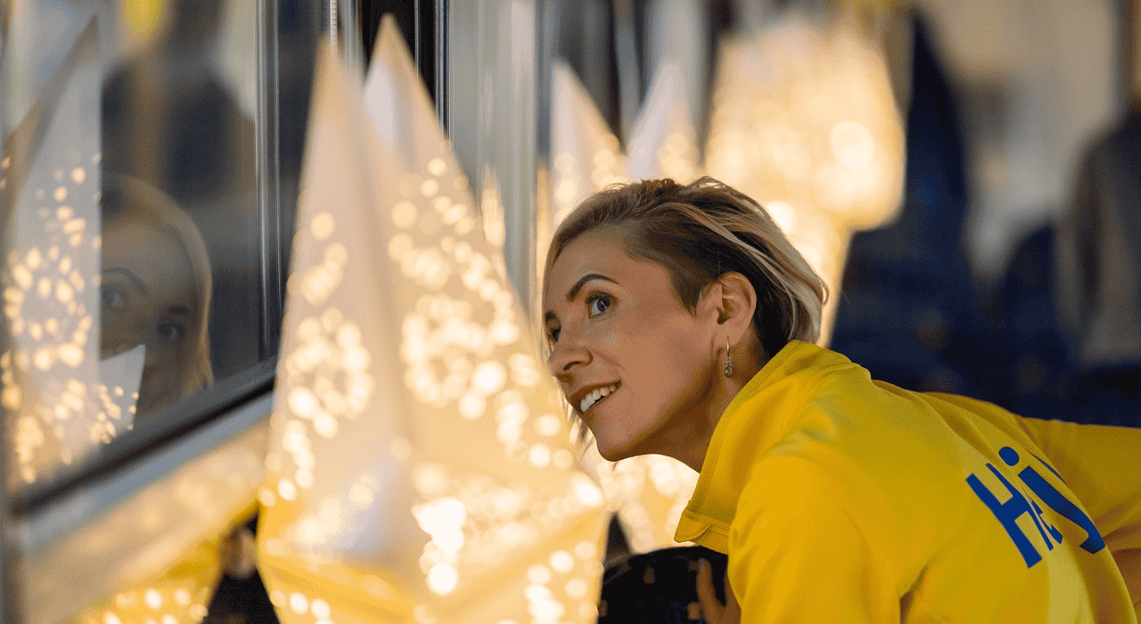 Sieviete IKEA uniformā, priecīgā izbrīnā skatās pa logu līdzās veselai rindai, pie sienas piekārtu, STRÅLA zvaigžņu.