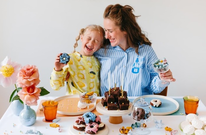 Мать и дочь в светлой комнате, украшенной яркими бумажными гирляндами. На белом столе — разноцветные блюда с кексами и пончиками, а также оранжевые стаканы и светло-голубые вазы с розовыми и оранжевыми цветами.