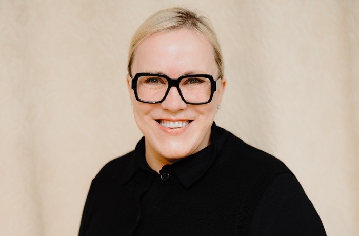 На бежевом фоне изображена улыбающаяся женщина со светлыми волосами, в черном свитере и черных очках.