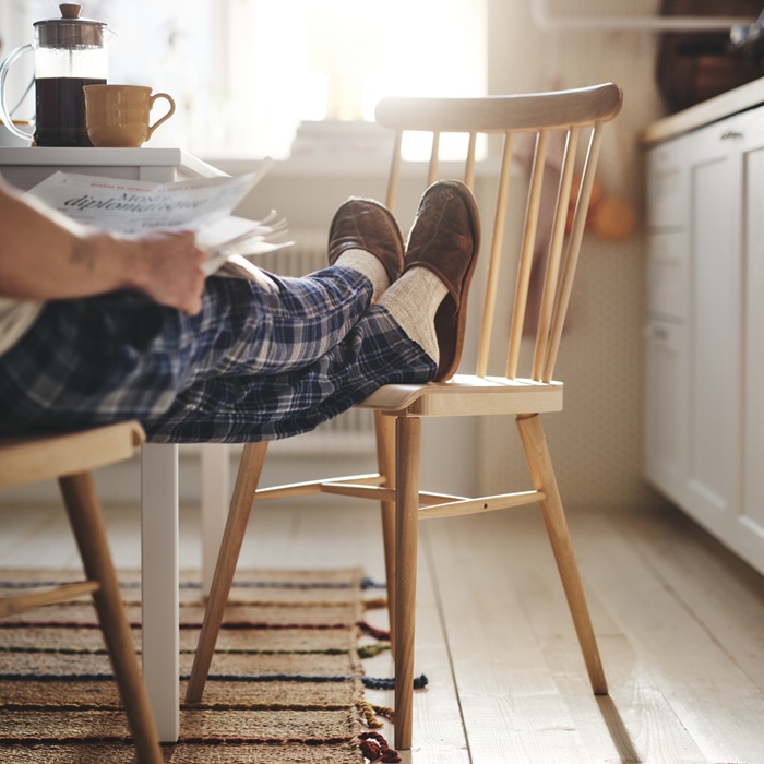 A person is sitting at the dining table with their feet, wearing slippers, resting on a wooden chair.