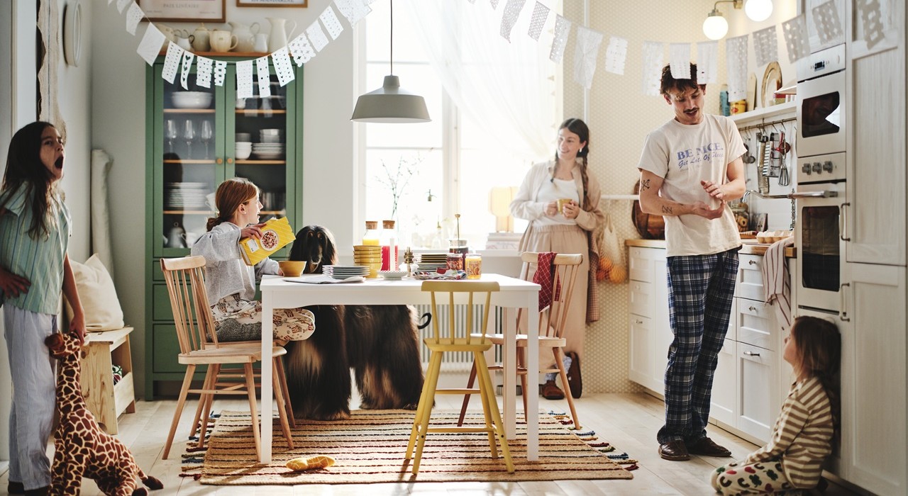 A bright, cosy kitchen with white cabinets, wooden chairs and a dining table, decorated with hanging garlands. Two adults, three kids and a dog are getting ready for a meal.