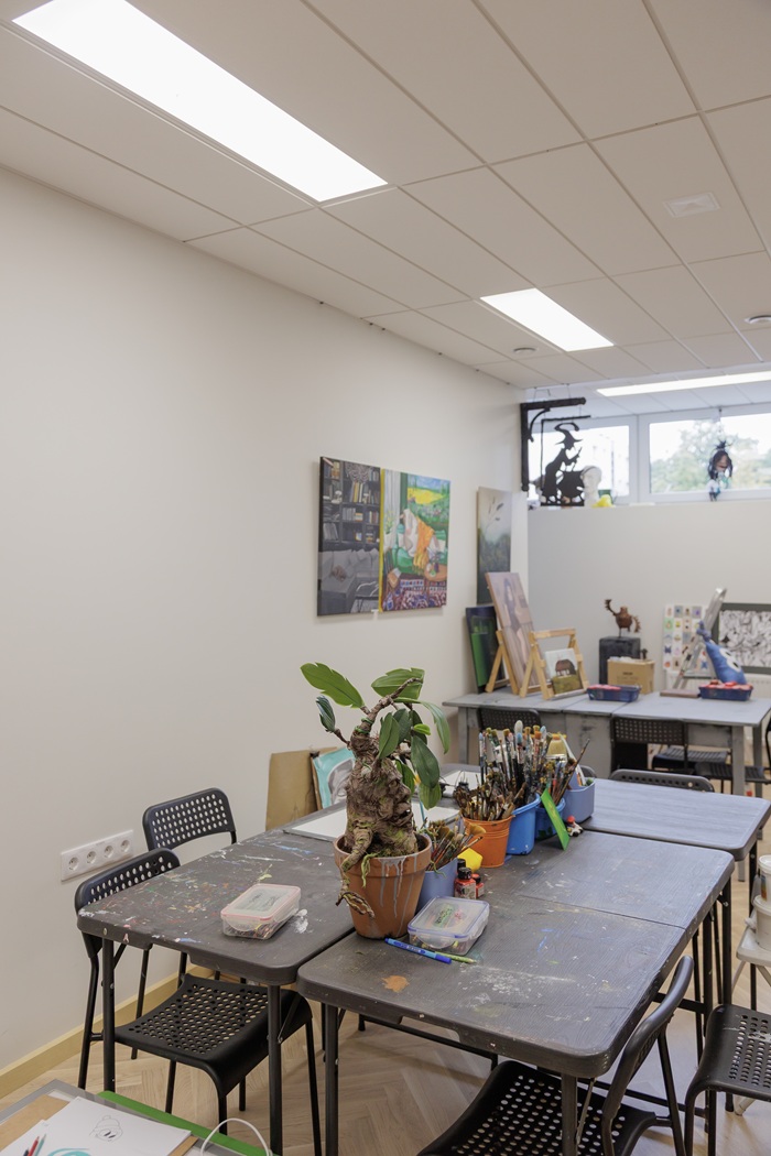 Dailės studija iki pokyčių: didelis stalas ir kėdės su akivaizdžiais nusidėvėjimo požymiais.