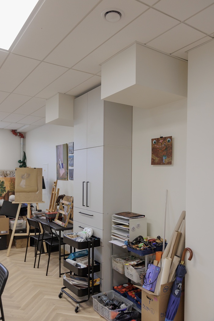 Darbo erdvė, pilna išmėtytų dailės reikmenų ir medžiagų, kurioms trūksta vietos.