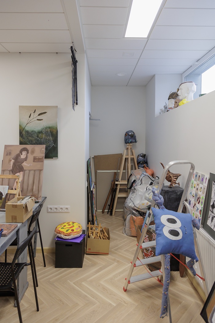 Atnaujinimo laukiančios studijos interjeras su netvarkingai išmėtytais baldais ir dailės reikmenimis.