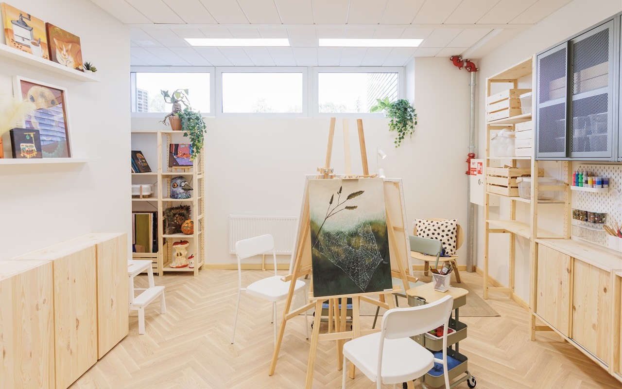 Šviesi ir tvarkinga dailės studija su medinėmis lentynomis, paveikslu ant molberto patalpos centre, kėdėmis ir tvarkingai išdėliotais dailės reikmenimis.