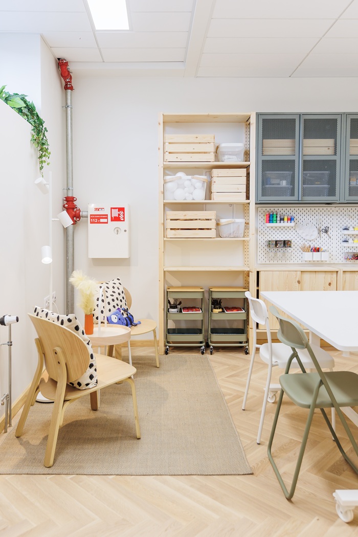 Šviesi dailės studija su medinėmis lentynomis, tvarkingai išdėstytais stalais ir kėdėmis.