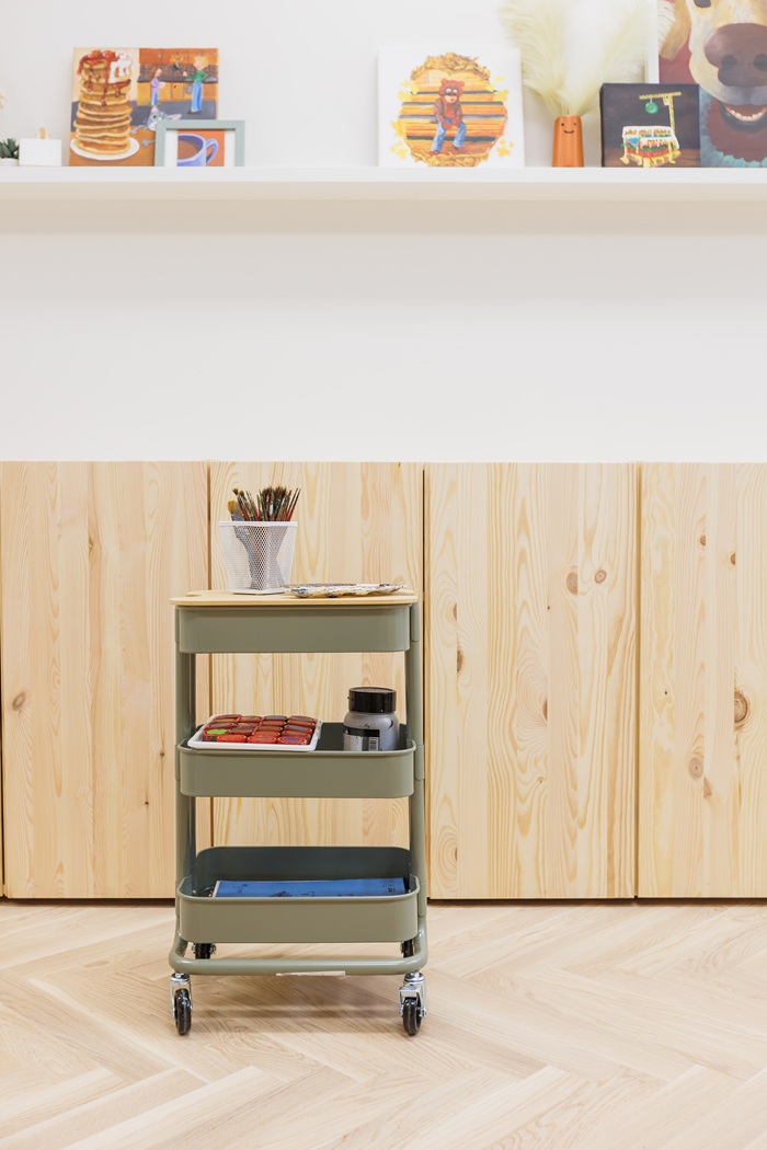 Priešais medines spinteles dailės studijoje stovi žalias vežimėlis su kūrybos priemonėmis.