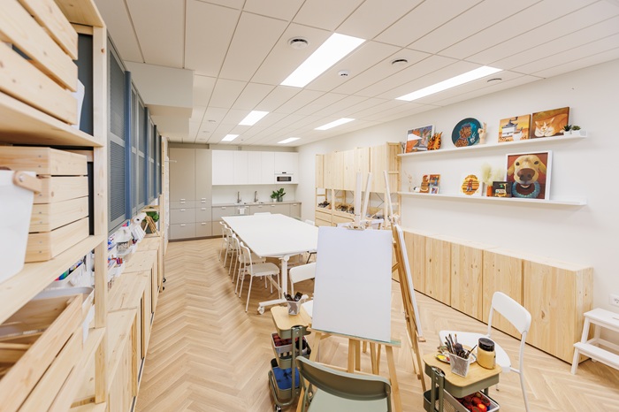 Šviesi dailės studija su medinėmis lentynomis, stalais ir molbertu, paruoštu kūrybiniam darbui.