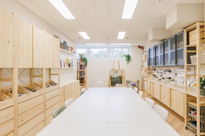 Erdvi dailės studija su mediniais daiktų laikymo baldais, ilgu darbo stalu, sudarytu iš kelių rašomųjų, ir kūrybai skirtų priemonių pilnomis lentynomis.