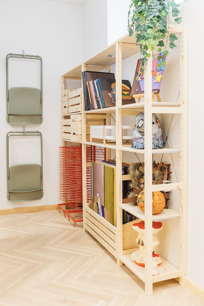 Atviros medinės lentynos, pripildytos knygų, dėžučių ir dailės reikmenų.