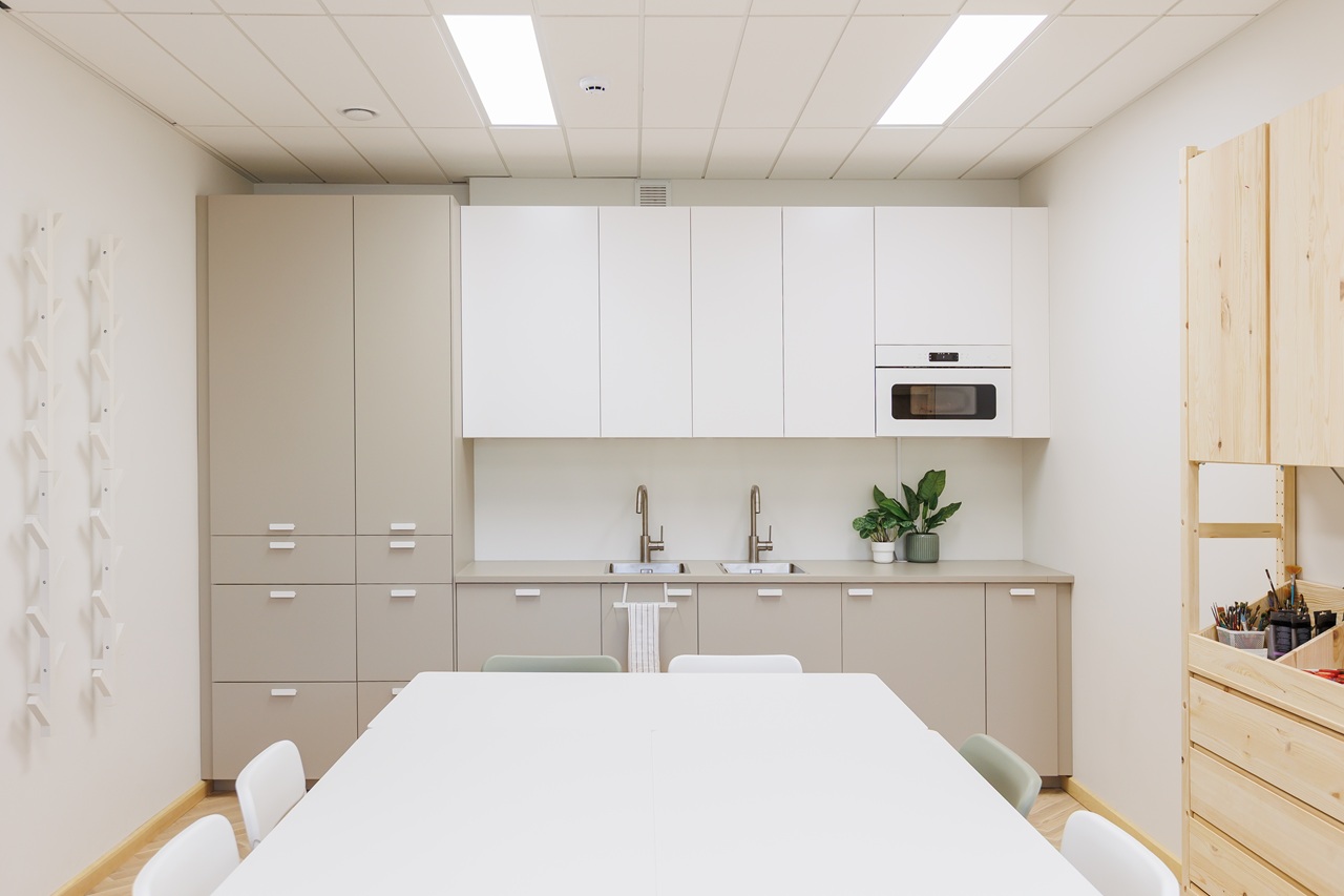 Šviesi, minimalistinė virtuvės zona dailės studijoje su smėlio spalvos spintelėmis, dviguba plautuve, dideliu baltu stalu ir dailės reikmenimis ant atvirų medinių lentynų.