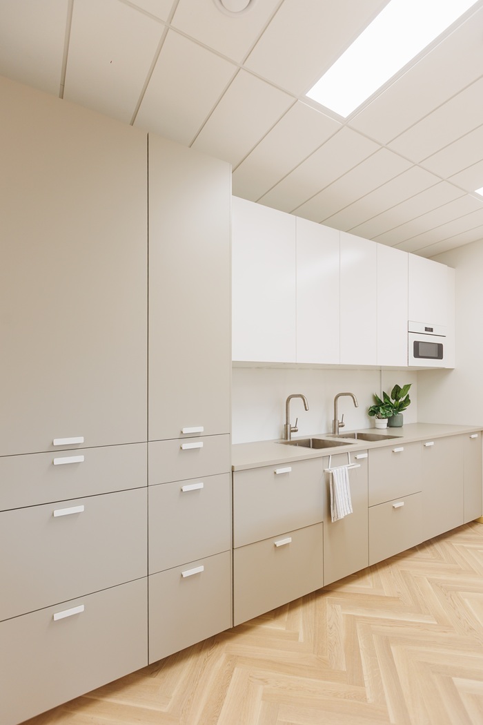 Minimalistinio stiliaus virtuvė vaizdas iš šono: spintelės su smėlinės spalvos fasadais, dviguba plautuvė ir nedidelis vazoninis augalas ant stalviršio.