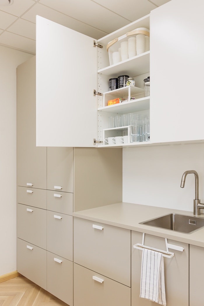 Virš plautuvės įrengta virtuvinė spintelė praviromis durimis: viduje matosi tvarkingai sudėti indai ir įstatomosios lentynėlės.