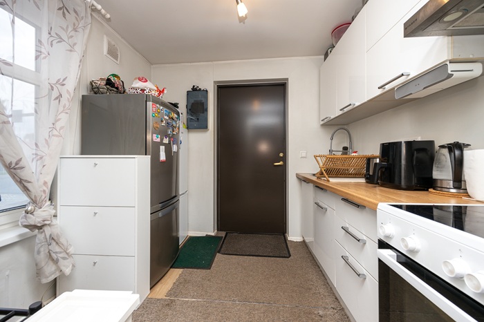 Vaade kitsasse kööki, mille parema seina ääres on valged köögikapid ja vasakus küljes kaks külmkappi. Ruumi kaugemas otsas on pruun uks.