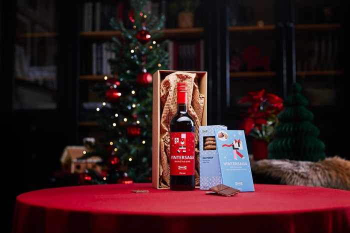 A festive table with a red tablecloth displays a gift set including a bottle of glögg, a box of milk chocolate and a package of oat biscuits. In the background, a decorated Christmas tree and holiday decorations create a warm atmosphere.