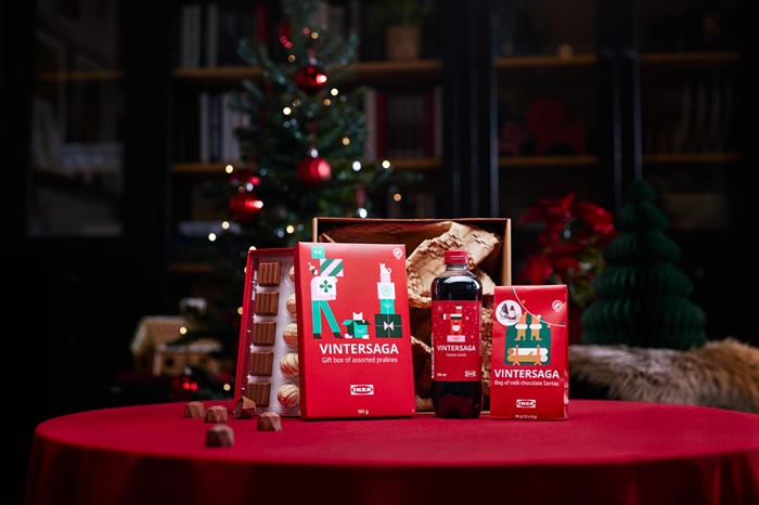 A festive table with a red tablecloth displays a gift set including a bag of chocolate Santas, a bottle of festive drink and a box of assorted pralines. In the background, a decorated Christmas tree and holiday decorations create a warm atmosphere.