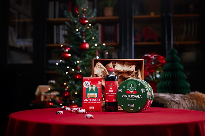 A festive table with a red tablecloth displays a gift set including a bag of chocolate Santas, a bottle of festive drink and a tin with ginger thins. In the background, a decorated Christmas tree and holiday decorations create a warm atmosphere.                                