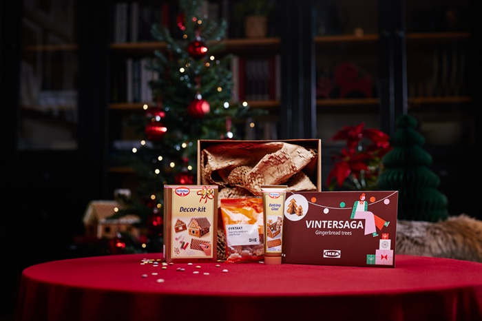 A festive table with a red tablecloth displays a gift set including a box of gingerbread trees, a pack of sour jelly candy, a decor kit and baking glue. In the background, a decorated Christmas tree and holiday decorations create a warm atmosphere.                                