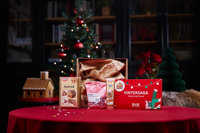 A festive table with a red tablecloth displays a gift set including a gingerbread house, a pack of jelly candy, a decor kit and baking glue. In the background, a decorated Christmas tree and holiday decorations create a warm atmosphere.                                
