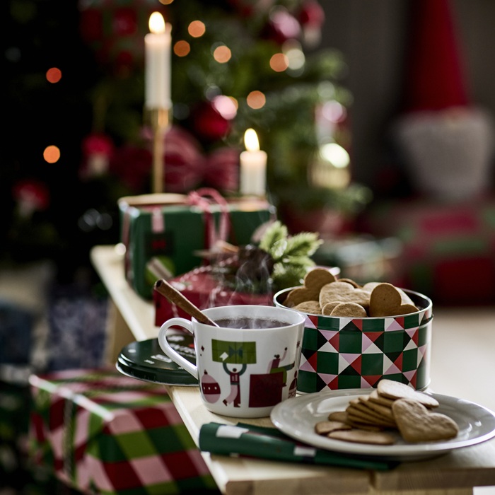A festive table setup with a cup of tea, a tin full of cookies and a plate with biscuits. The table is decorated with red, green and white patterns, matching wrapped gifts nearby.