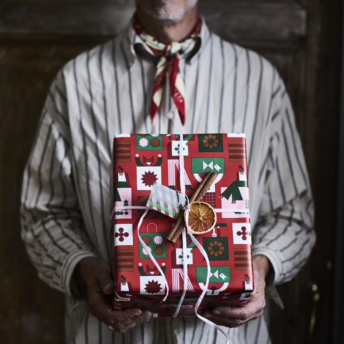 A person wearing a striped shirt and scarf holds a gift wrapped in festive red, green and white paper, decorated with ribbon, a dried orange slice and a cinnamon stick.