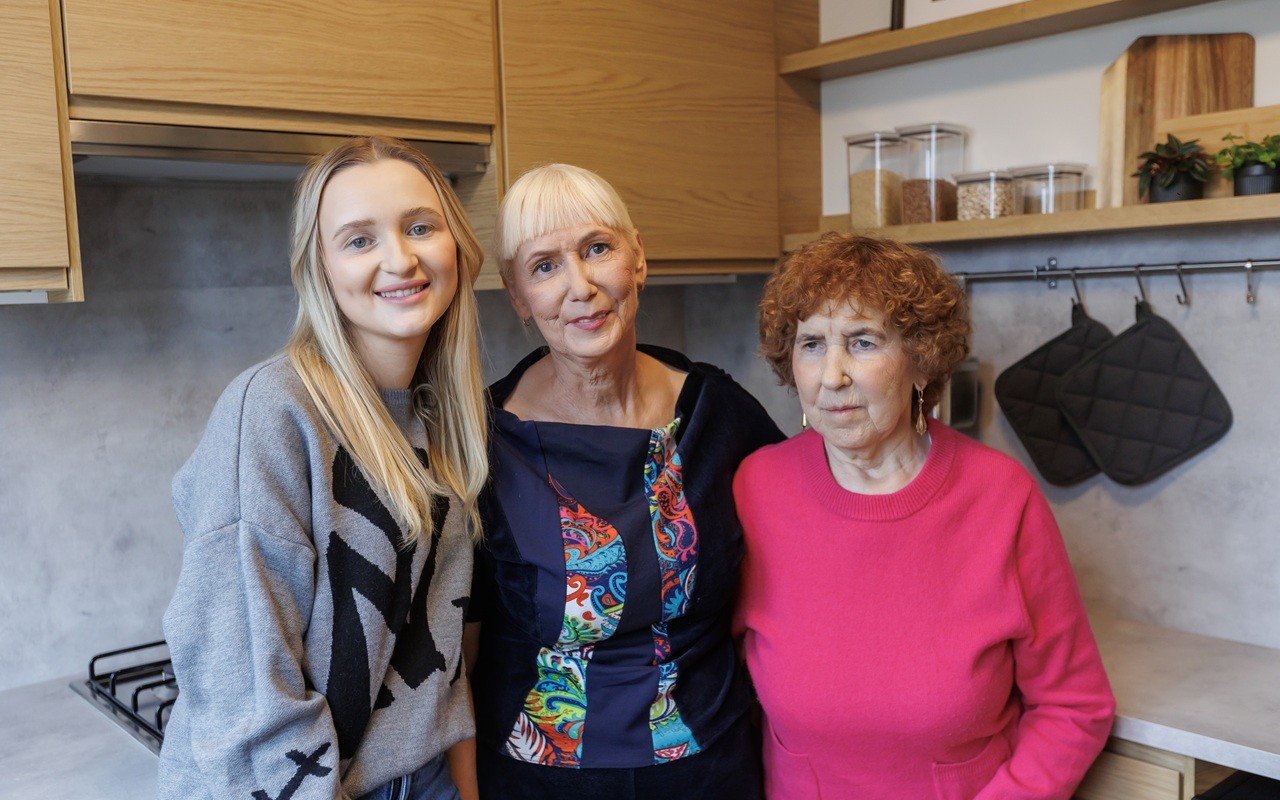 Trijų kartų atstovės – dukra, mama ir močiutė – stovi modernioje virtuvėje ir šypsosi.