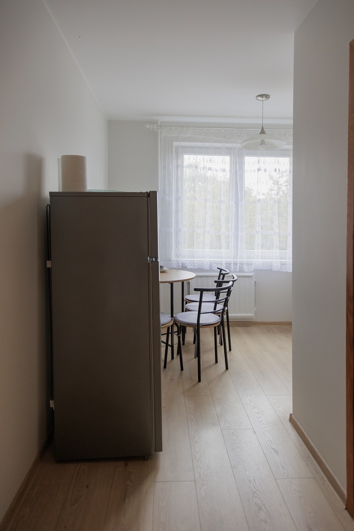 Virtuvės vaizdas iki pokyčių: siaura erdvė su šviesios medienos grindimis, šaldytuvu ir nedideliu valgomojo stalu prie lango.
