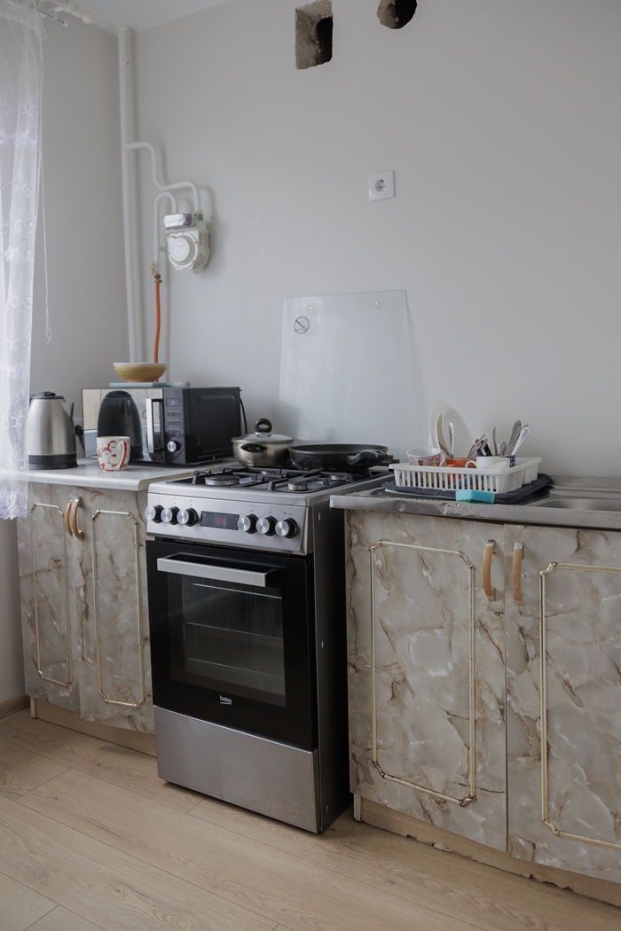 Virtuvės vaizdas iki pokyčių: paprastai įrengta virtuvė su sena virykle, prietaisais ant stalviršio ir minimaliu dekoru.