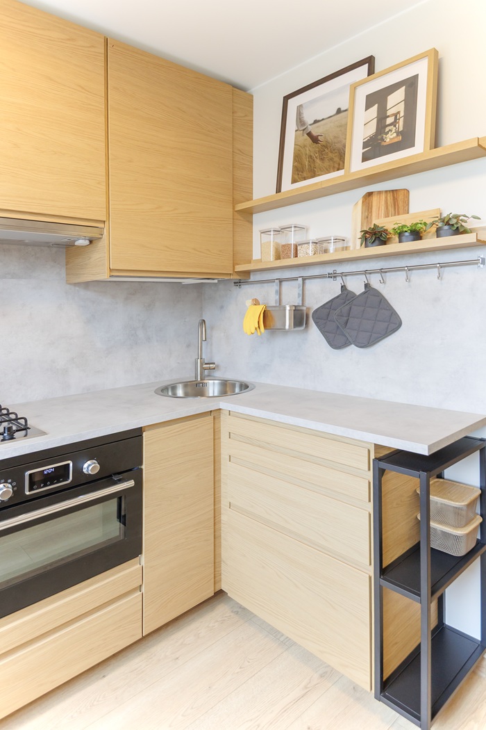 Kompaktiška virtuvė su šviesios medienos fasadais, stalčiais ir atviromis lentynomis, padedančiomis maksimaliai išnaudoti erdvę ir palaikyti tvarką.