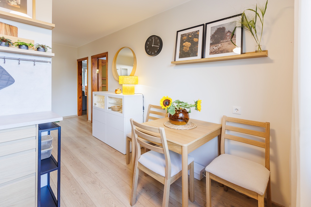 Modernioje virtuvėje įrengta valgomojo zona su mediniu stalu, dviem kėdėmis, vazoje pamerktomis saulėgrąžomis ir įrėmintomis nuotraukomis ant sieninės lentynos.